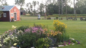 A small red building with a door and windows, surrounded by a well-maintained garden with various flowers and a windmill in the background. - Garden Lodging In A Peaceful Country Farm Setting ~ "Longspur" House  (Carrington)