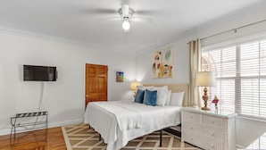 A bedroom with a bed, a ceiling fan, a television mounted on the wall, a wooden door, a window with curtains, and a bedside table with a lamp.