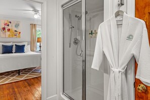 A hotel room with a glass-enclosed shower, a white bathrobe hanging on the door, a bed with blue pillows, and a colorful abstract painting on the wall. - Charlotte at Jubilee Suites Bed & Breakfast. 1K/1B (Fairhope)