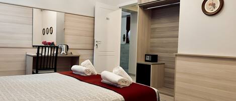 A hotel room with a bed, a desk, a chair, a mirror, a door, and a small safe.