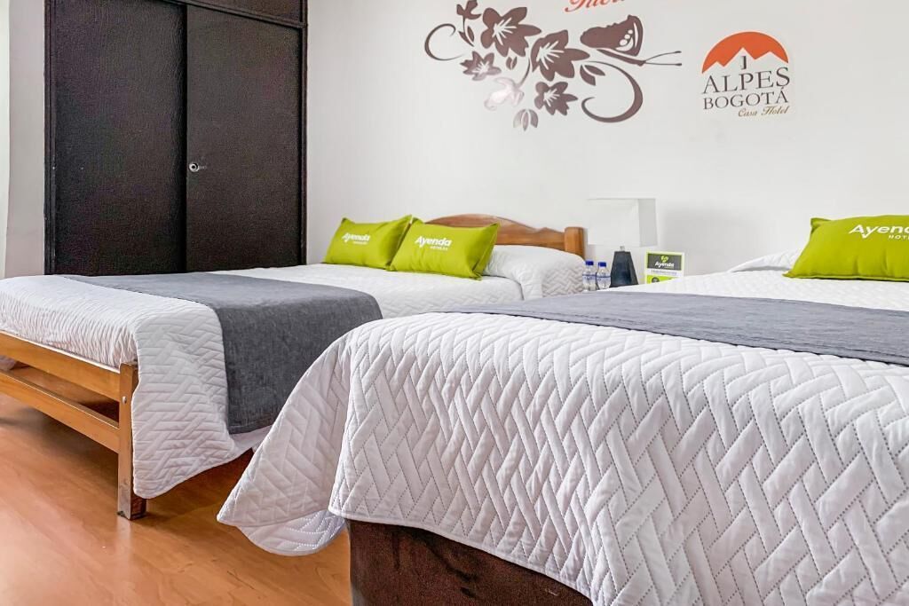 A hotel room with two beds, each with white bedding and green pillows, a wooden wardrobe, and a wall decoration for 'Alpes Bogotá Casa Hotel'.