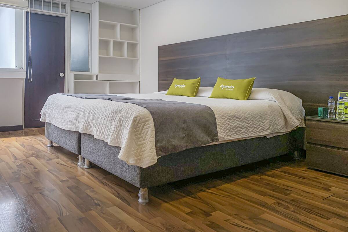 Una cama bien tendida con cabecera gris y dos almohadas verdes, suelo de madera y pared blanca con una estantería empotrada.