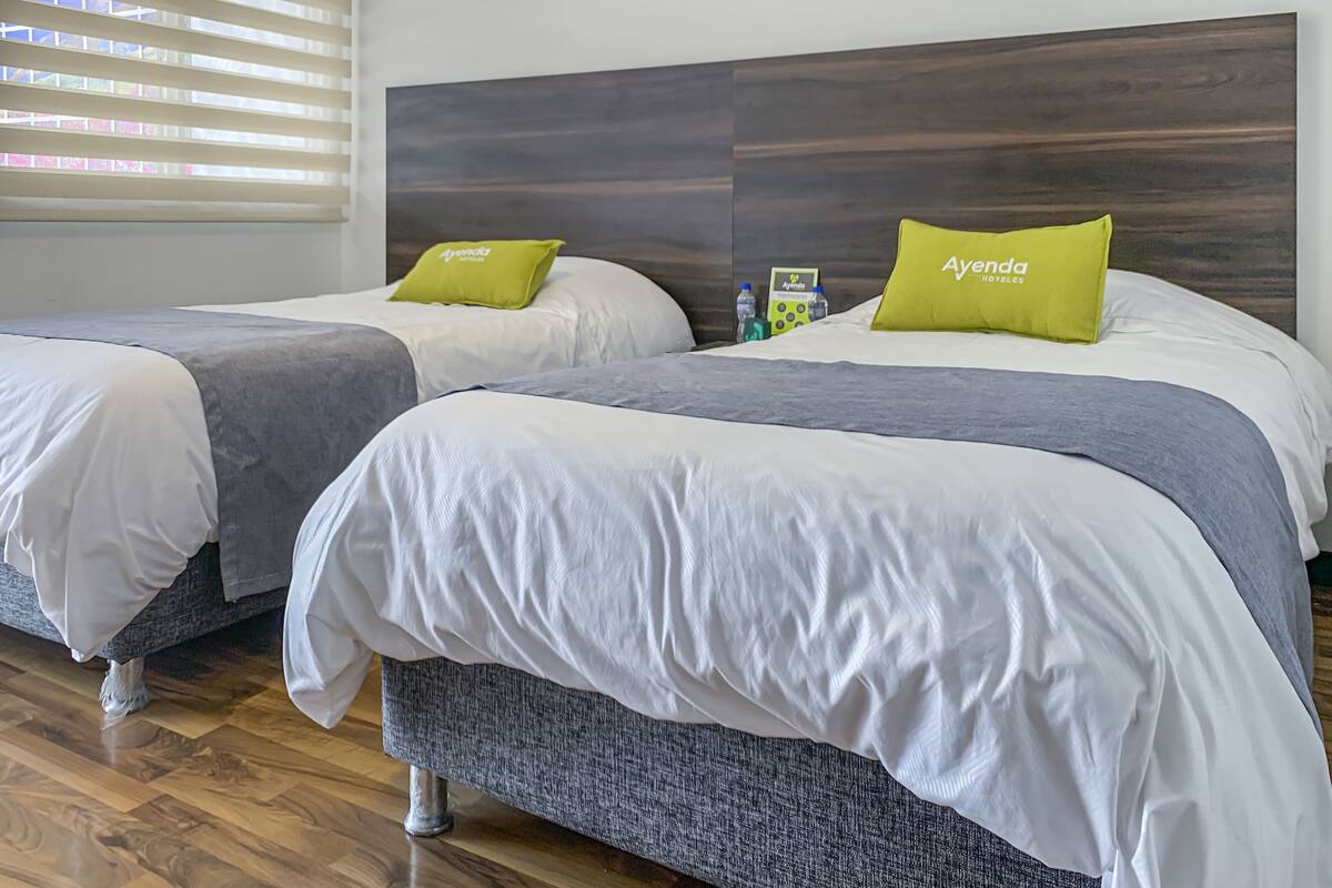 Una habitación de hotel con dos camas, cada una con una colcha gris y un almohadón verde con la etiqueta 'Ayenda Hotels'.