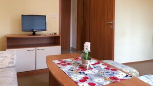 A room with a wooden desk, a television on a stand, a door, and a small table with a floral tablecloth. - Hotel Beatris (Nessebar)