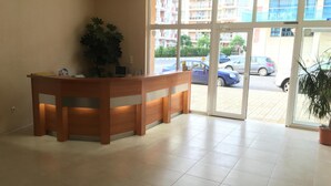 A reception desk with a potted plant, a tiled floor, and a view of parked cars and buildings through large glass doors. - Hotel Beatris (Nessebar)