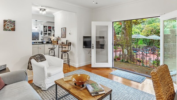 Amplia sala de estar con una mesa de centro de madera, un sillón blanco y un sofá. Se ve una cocina al fondo, y una puerta corrediza de vidrio da a un patio exterior con vistas a zonas verdes.