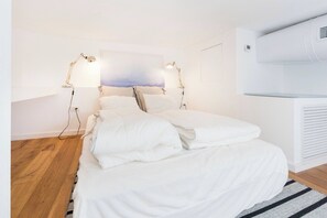 A bedroom with a bed, two bedside lamps, a wall-mounted air conditioner, and a wooden floor.