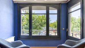 Ein Zimmer mit einem großen Erkerfenster, zwei grauen Sesseln und Blick auf eine Straße mit Autos und Bäumen.