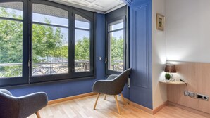 Ein Zimmer mit einem großen Fenster, einem Sessel, einem kleinen Tisch mit einer Lampe und einem Wandregal.