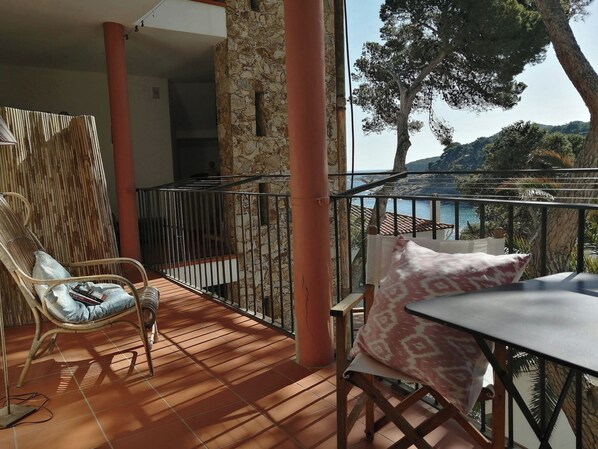 A balcony with a view of the sea, a wicker chair, a table with a cushion, and a stone pillar.