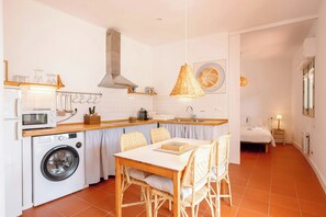 A compact kitchen with a washing machine, a dining table with chairs, a hanging light fixture, and a bed in the adjacent room.