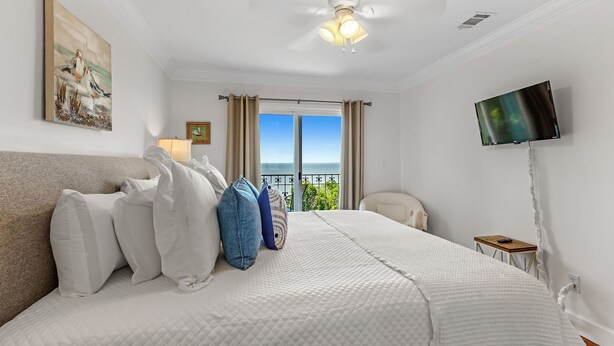 A bedroom with a large bed, a ceiling fan, a TV mounted on the wall, and a view of the sea through the window. - Cypress at Jubilee Suites Bed & Breakfast. 1K/1Q/1B (Fairhope)