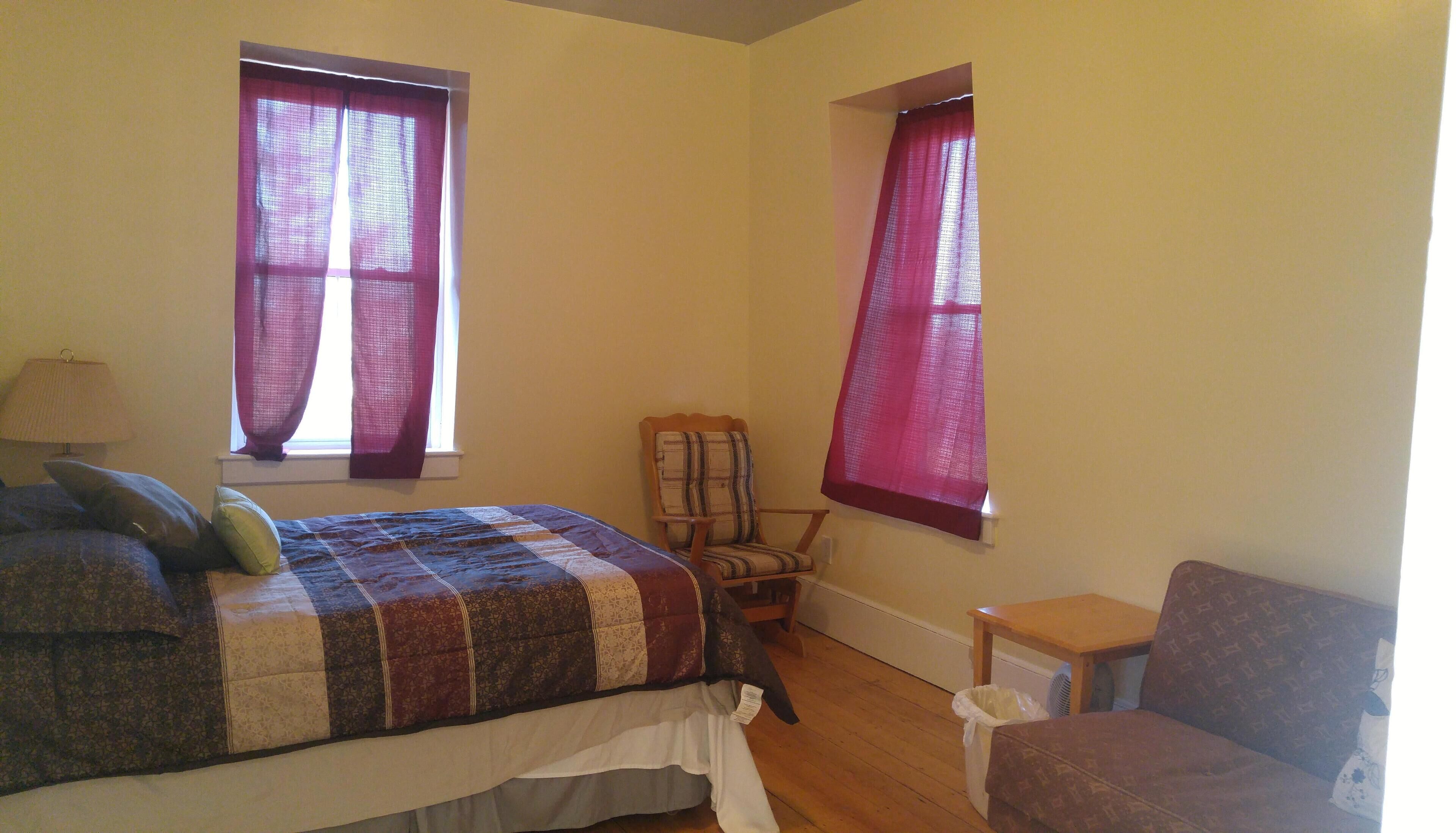 A bedroom with a bed, a chair, a small table, and two windows with curtains.