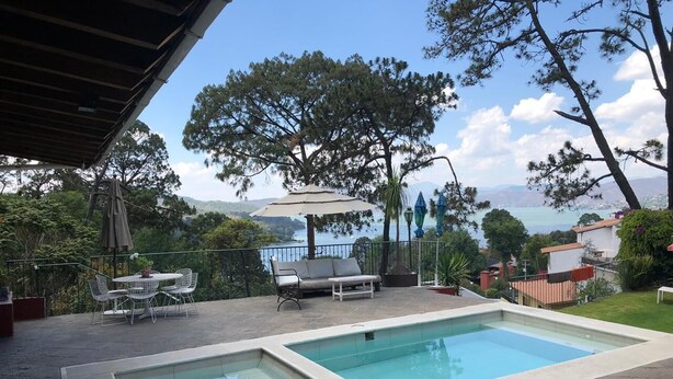 Una piscina rectangular con borde blanco, rodeada de baldosas de piedra. Hay una zona de estar cubierta con una mesa y sillas, y un gran paraguas.