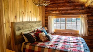 Quarto de cabana de madeira com uma cama, uma janela com cortinas e um lustre.