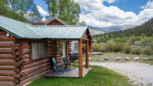 Uma cabana de toras com varanda, um carro e vista para montanhas e floresta.