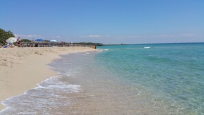 Ein Strand mit klarem türkisblauem Wasser, einem Sandstrand und einem Holzsteg mit Sonnenschirmen.