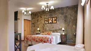 A hotel room with a large bathtub, two beds with floral bedding, a stone wall, and a chandelier. - Posada del Sauce (Villa General Belgrano)