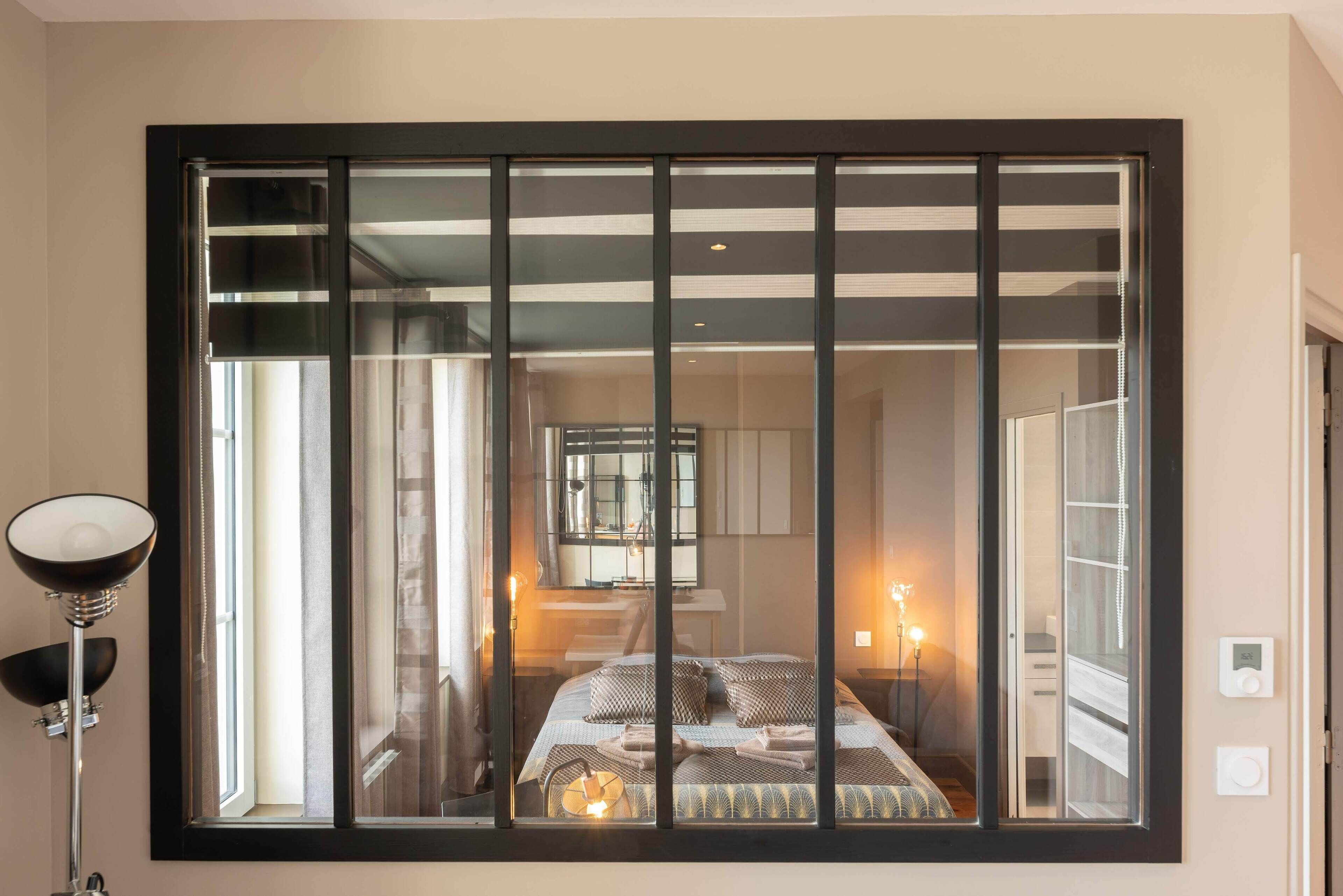 A bedroom with a large window, a bedside lamp, and a bed with pillows.