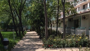 Un sendero pavimentado bordeado de árboles y macizos de flores, que conduce a un edificio con balcones.