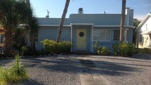 A two-story house with a yellow front door, surrounded by gravel and palm trees.