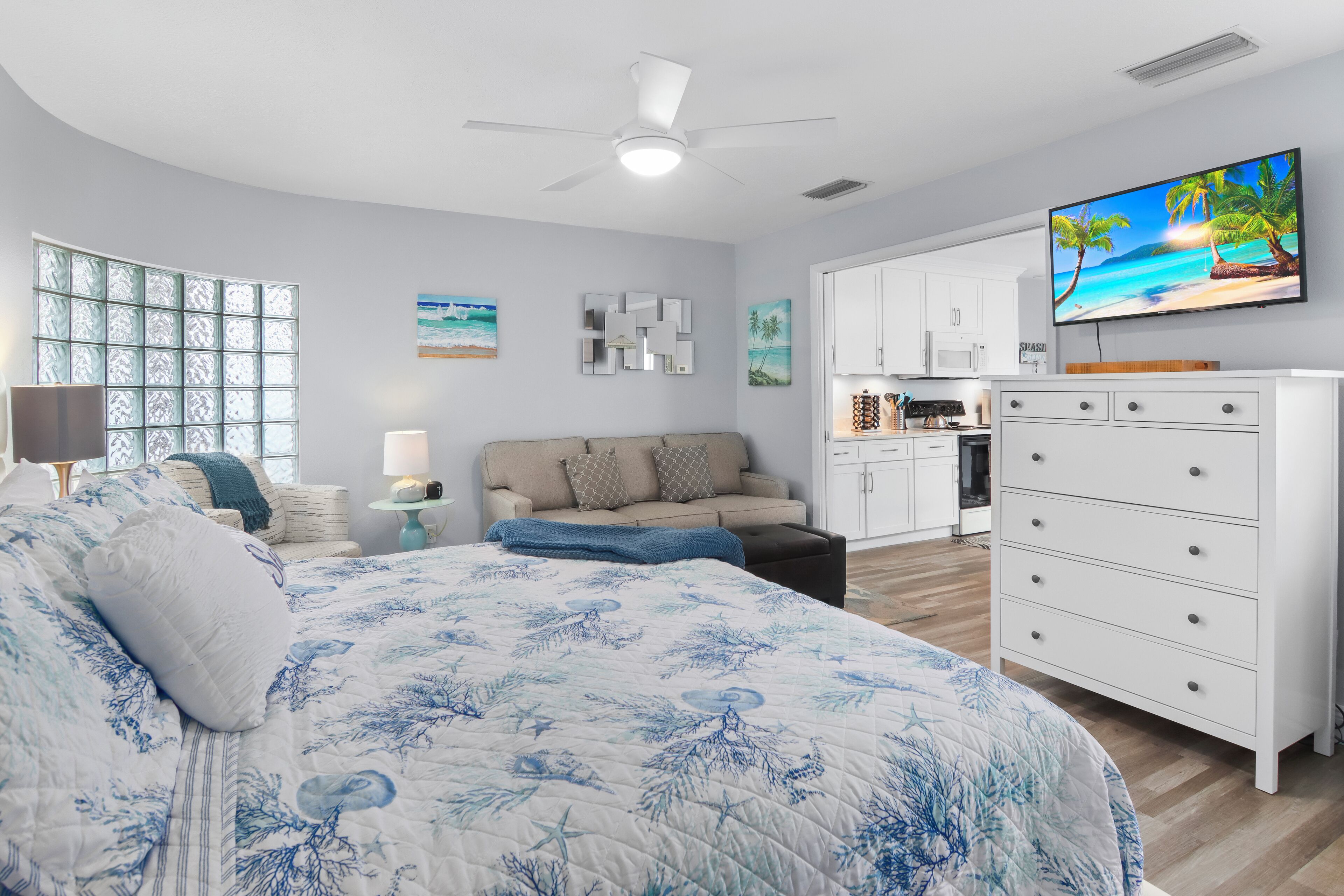 A bedroom with a large bed, a TV, a kitchenette, and a view of the beach.