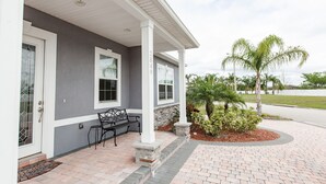 A house with a covered porch, a stone column, a bench, and a small garden with palm trees.