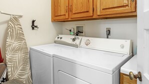 A laundry area with a front-loading washer and dryer, wooden cabinets above, and a towel rack with a leaf-patterned towel. - Magnolia at Jubilee Suites Bed & Breakfast. 1K/1B. (Fairhope)