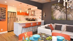 Une cuisine moderne dotée d’un îlot central, de tabourets de bar orange et d’une table à manger agrémentée d’assiettes et de verres bleus.