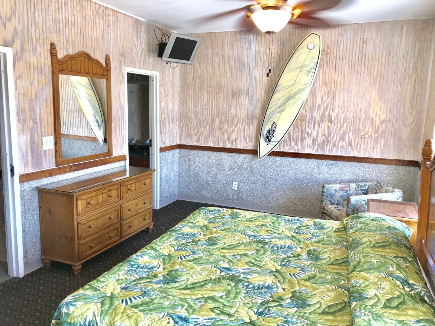 A bedroom with a bed, a dresser, a mirror, a television, and a surfboard decoration.
