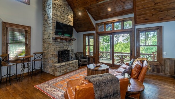 A spacious living room with a stone fireplace, wooden ceiling, large windows, and a cozy seating area. - Prancing Horse at Eagles Nest - hot tub, great view, resort amenities, summer concerts! (Banner Elk)