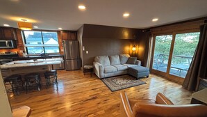 A modern living space with a kitchen, a grey sectional sofa, a brown leather chair, and a large window offering a view of the outdoors.