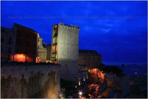 Una torre de piedra histórica con balcón, rodeada de edificios con ventanas iluminadas, mostrando un paisaje urbano nocturno.