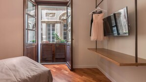 A bedroom with a bed, a TV mounted on the wall, a wooden shelf, and a door leading to a balcony with a view of a building. - Suite 101 - Cagliari (Cagliari)