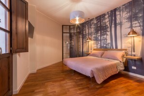 Un dormitorio con piso de madera, una cama con colcha rosa, un televisor montado en la pared y dos lámparas también fijadas a la pared.