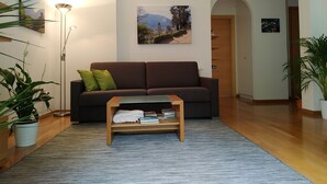 Uma sala de estar com um sofá marrom, almofadas verdes, uma mesa de centro de madeira e um quadro decorativo de uma paisagem na parede.