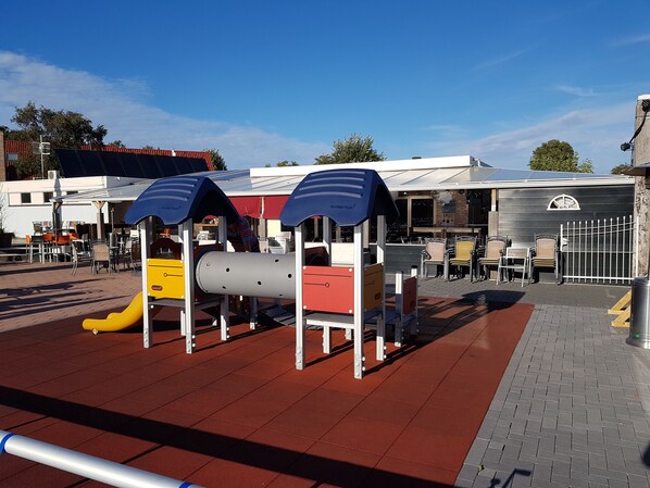 A playground with slides and play structures, surrounded by outdoor seating and a building with a red roof. - Mobile Home in Renesse at the 4-star Camping de Brem (Renesse)