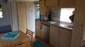 A compact kitchen with a sink, stove, and cabinets. There is a dining table with chairs and a window providing natural light. - Mobile Home in Renesse at the 4-star Camping de Brem (Renesse)