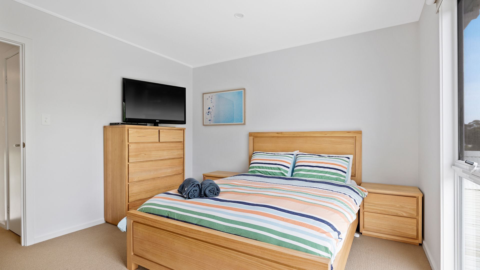 A bedroom with a bed, a TV on a stand, a nightstand, a wooden dresser, and a window.