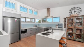 Eine moderne Küche mit einem Edelstahl-Kühlschrank, einem eingebauten Ofen, einem Gasherd und einer großen Kochinsel mit Hockern. Mehrere Fenster sorgen für natürliches Licht und einen Ausblick ins Freie.