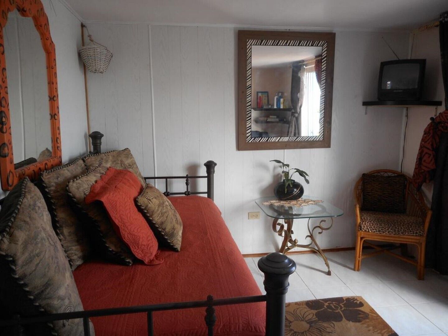 A bedroom with a bed, a chair, a small table with a plant, a mirror, and a television on a shelf.