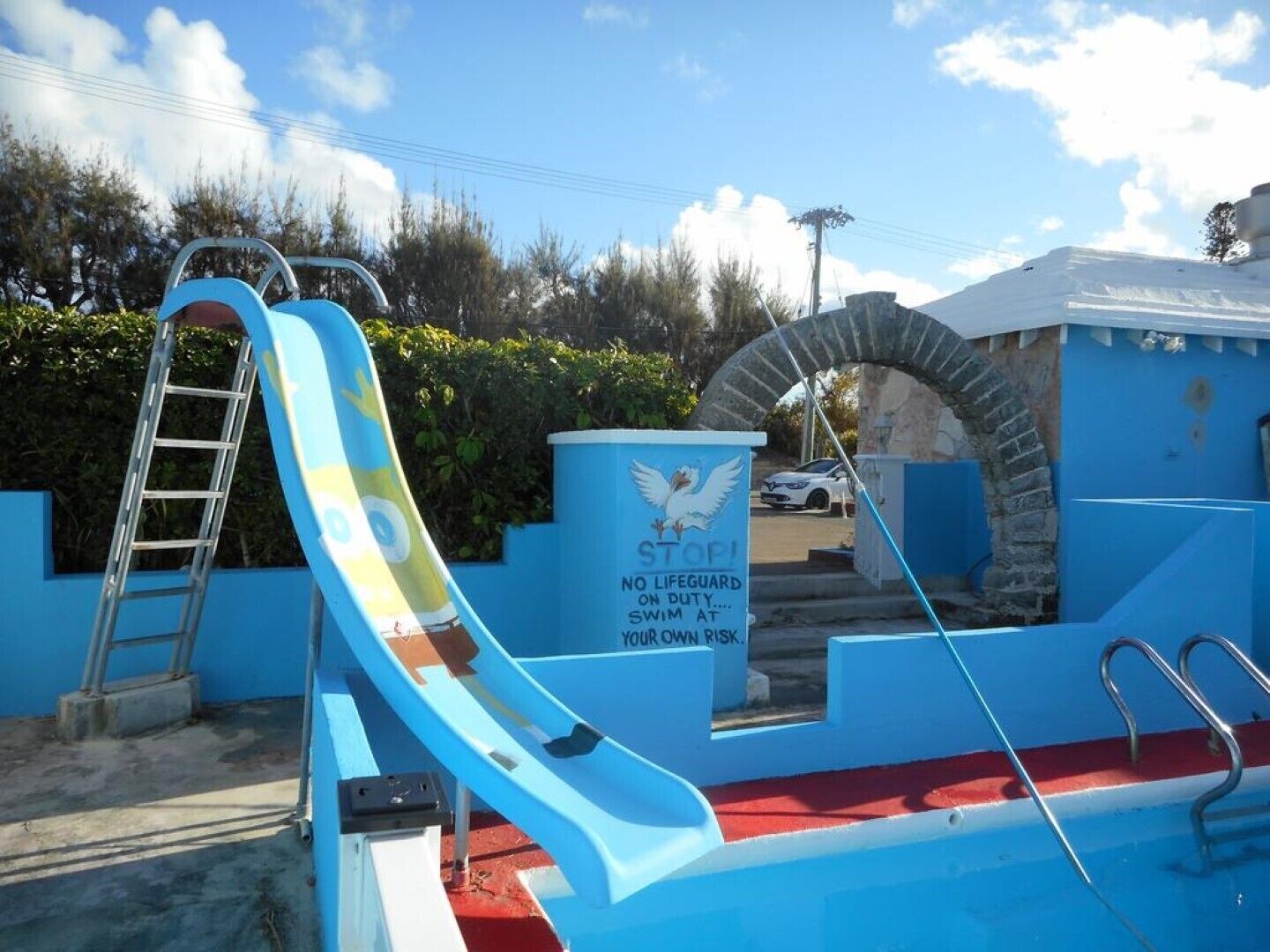 A swimming pool with a blue slide, a lifeguard sign, and a stone archway.