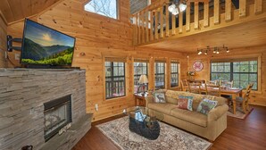 A cozy cabin interior with a stone fireplace, a television mounted on the wall, a dining area with a table and chairs, and large windows offering views of the outdoors. - Best Price!!! Cabin with Mountain Views, Hot Tub, Sleeps 6, 10 Min to Parkway! (Sevierville)