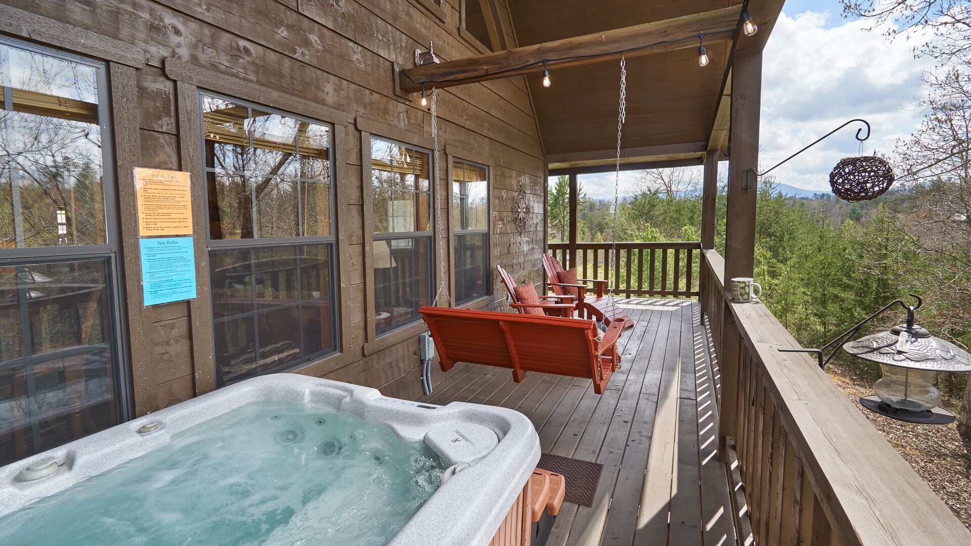 Una terraza de madera con un jacuzzi, sillas Adirondack rojas y vistas a árboles y montañas.