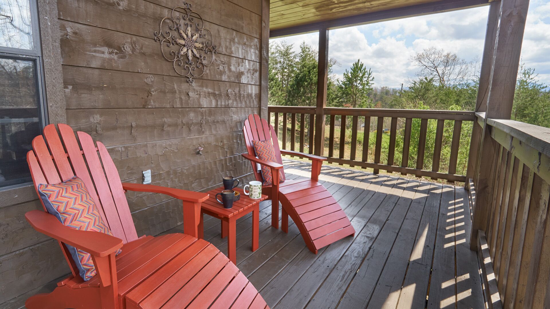 A wooden deck with two red Adirondack chairs, a small table with a mug and a cup, and a decorative metal piece on the wall.