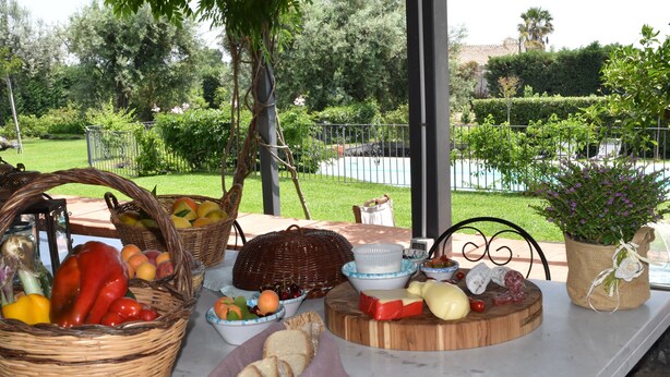 An outdoor dining area with a table set with various food items, surrounded by greenery and a view of a pool. - Luxury Villa with Large Pool, Vineyard, Views of the Sea, Taormina  and Mt.Etna (Acireale)