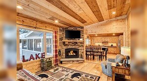 A cozy cabin interior with a fireplace, a television, a dining area, and a large window with the word 'LAKE' visible.