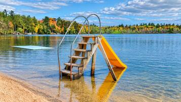 A wooden slide with metal supports in a lake setting, surrounded by trees and clear skies.