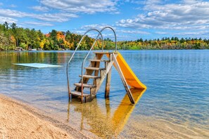 Un toboggan en bois, soutenu par des éléments métalliques, installé au bord d’un lac, entouré d’arbres et sous un ciel dégagé.