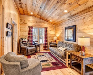 L’intérieur d’un chalet accueillant, avec des murs en bois, un canapé, deux fauteuils, une table et une fenêtre avec des rideaux.